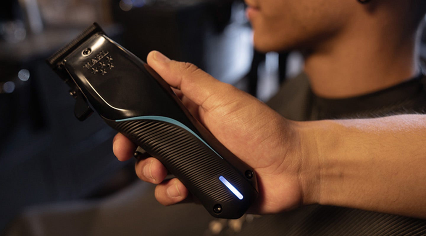 A hand holds a black hair clipper with a blue accent, ready for use on a person in a barber setting.