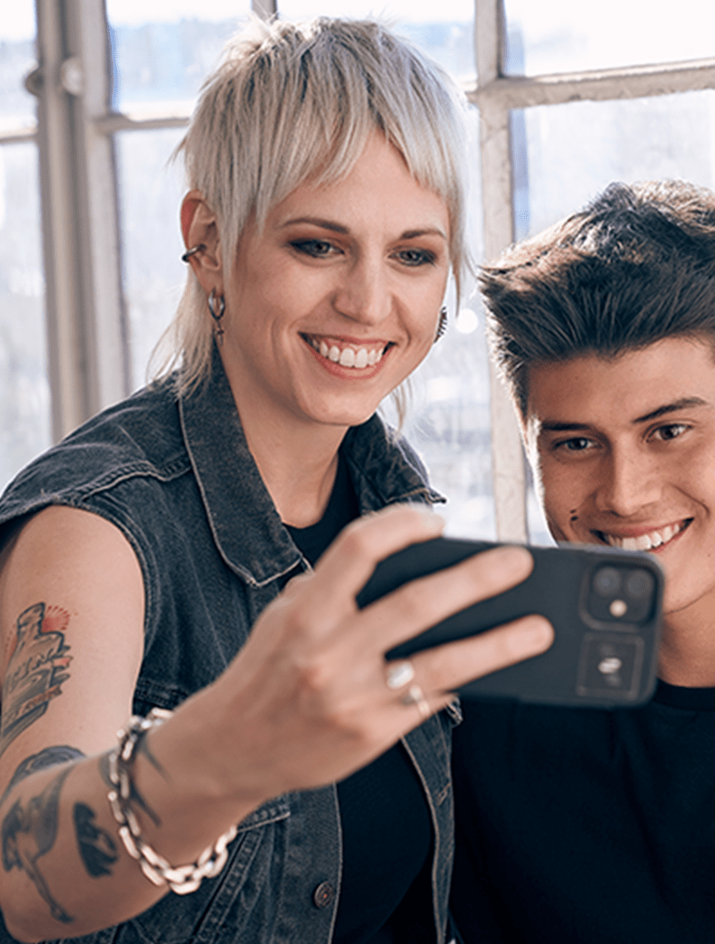 Female barber with long blonde hair dressed in a denim vest holding a phone posing for a selfie