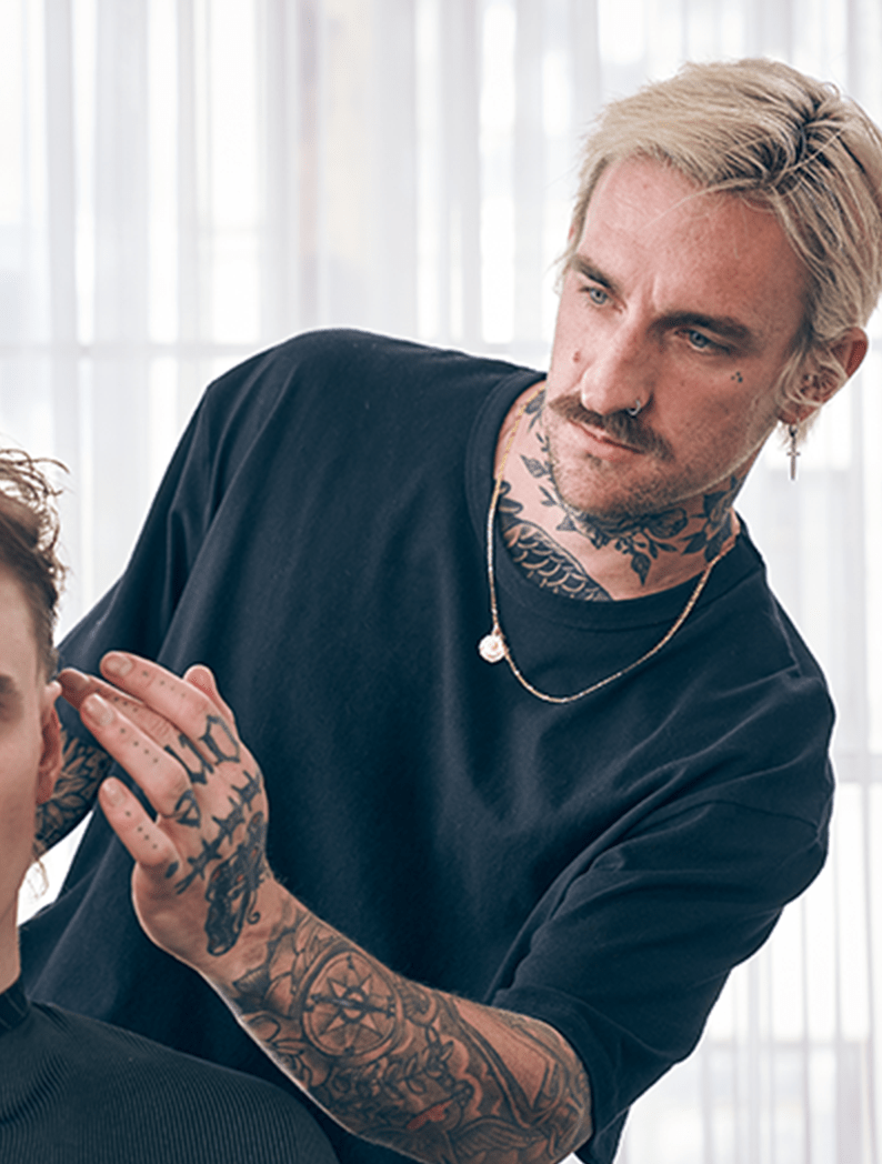 Male barber with long blonde hair and tattoos dressed in a black shirt styling hair