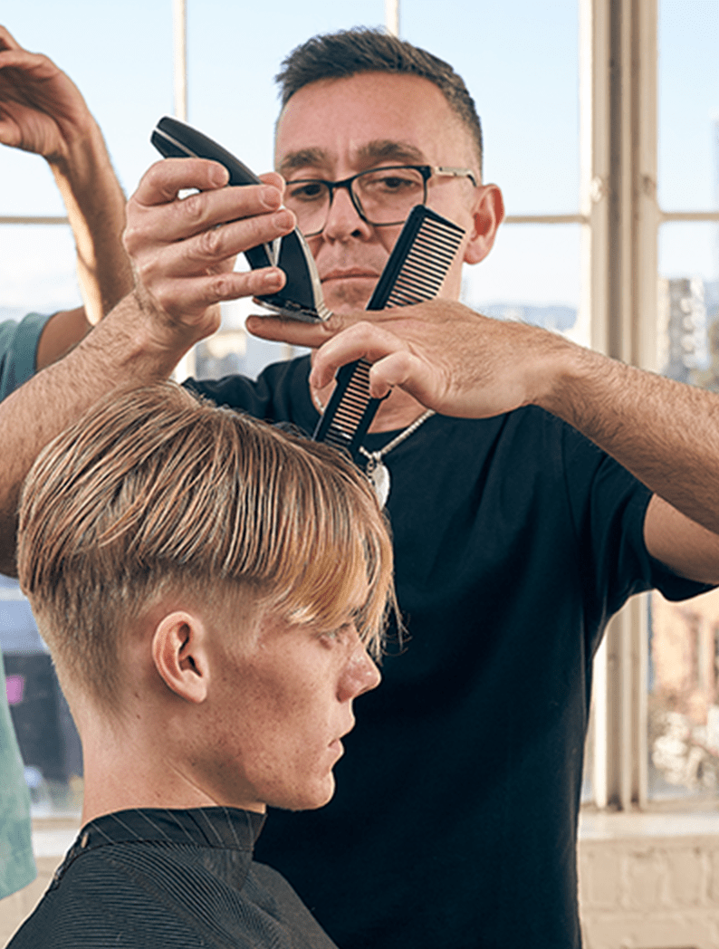 Male barber with short hair and glasses dressed in a black t-shirt holding a clipper and comb