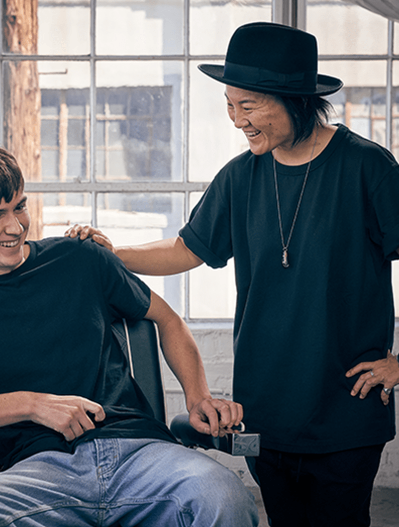 Female barber dressed in a black t-shirt and black fedora hat laughing with a model